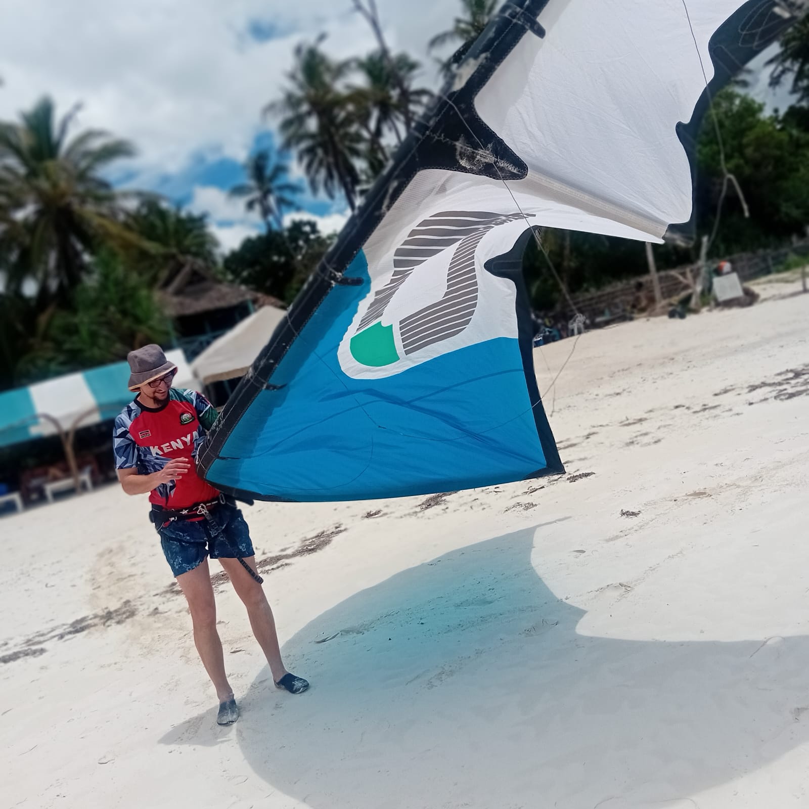 Instructor carrying a large blue kite along the beach with palm trees behind